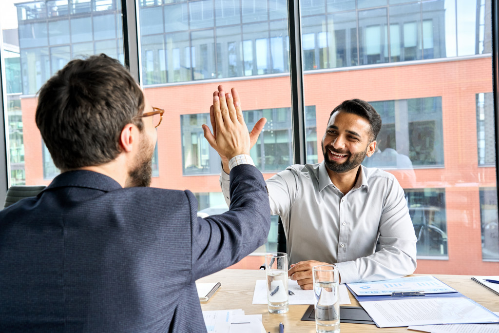 Two happy smiling diverse professional businessmen executive leaders giving highfive after successful financial business deal acquisition merger at board office meeting.