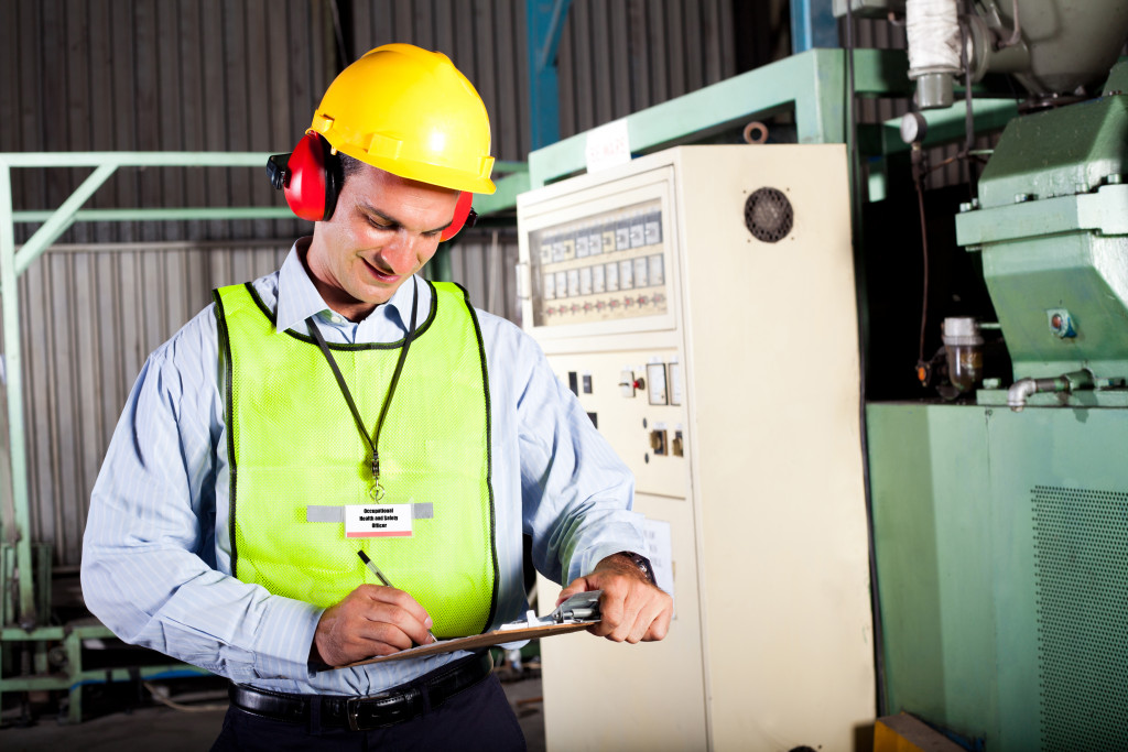 male inspector writing on clipboard while wearing hard hat, ear muffs, and reflective vest